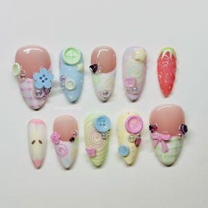 Puede incluir: Un conjunto de diez uñas postizas con diversos diseños en tonos pastel, que incluyen botones, espirales, una fresa, un lazo y patrones de cuadros. Las uñas están decoradas con pequeñas cuentas y dijes, con el texto pressonhaven visible en el centro.