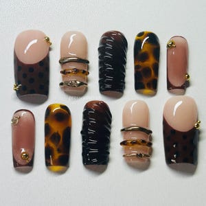 Könnte beinhalten: Eine Kollektion von zehn künstlichen Fingernägeln in verschiedenen Designs. Einige Nägel haben eine nackte Basis mit braunen Punkten oder eine braune Spitze. Andere haben ein Schildpatt- oder strukturiertes braunes Design. Goldakzente sind vorhanden.