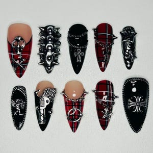 Puede incluir: Un juego de diez uñas postizas puntiagudas con una mezcla de patrones de cuadros rojos y negros, superficies negras sólidas y adornos de metal plateado que incluyen calaveras, cruces, cadenas, pinchos, estrellas y un revólver.