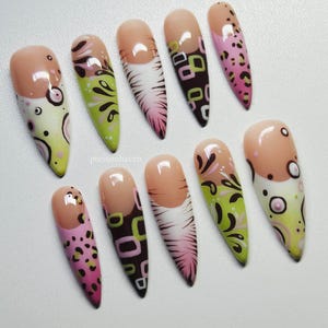 Puede incluir: Un conjunto de diez u&ntilde;as postizas largas en forma de almendra. Cada u&ntilde;a presenta una base nude y varios dise&ntilde;os, incluyendo estampado de leopardo rosa, espirales verdes y negras y patrones geom&eacute;tricos en marr&oacute;n y verde. Las u&ntilde;as est&aacute;n dispuestas sobre una superficie blanca.