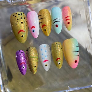Puede incluir: Un juego de diez uñas postizas en forma de almendra dispuestas en un estuche de plástico transparente. Cada uña presenta un diseño inspirado en peces con colores vibrantes, acabados con purpurina, ojos pintados y detalles de boca.