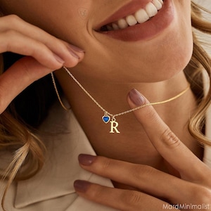 以下が含まれることがあります： 小さな青いハート型のペンダントと文字「R」が付いたゴールドカラーのネックレス。チェーンは細く、ペンダントは小さいです。画像はネックレスに焦点を当てており、背景はぼやけています。