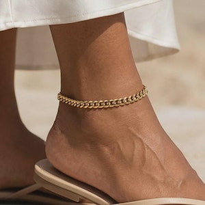 May include: A gold-coloured chain anklet. The anklet features a curb chain design and is worn on the ankle. The image shows the anklet on a person's foot, with a white garment in the background.