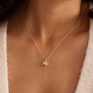 May include: A delicate gold necklace with a small pendant in the shape of the letter "A". The necklace is worn around the neck. The image is a close-up shot of the necklace and the person wearing it.