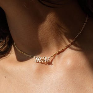 Puede incluir: Un collar de cadena dorada con las letras "RM" en cursiva. Las letras están adornadas con pequeñas piedras brillantes. El collar se lleva alrededor del cuello, con la cadena descansando sobre la piel.