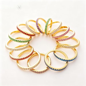 Op de afbeelding: Een verzameling goudkleurige ringen, elk versierd met een band van kleurrijke edelstenen. De ringen hebben een verscheidenheid aan kleuren, waaronder rood, groen, paars, roze, blauw en helder, waardoor een levendige weergave ontstaat.