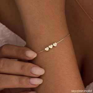 Pode incluir: Uma delicada pulseira dourada com três pingentes em forma de coração, cada um com uma inicial. A pulseira é usada no pulso, mostrando a joia personalizada. O design é minimalista e elegante.
