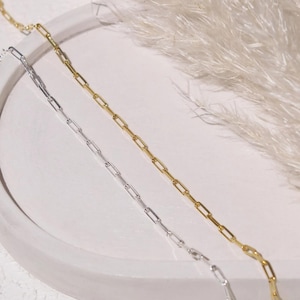 以下が含まれることがあります： 白い表面に、金と銀の2つの繊細なチェーンネックレスが表示されています。チェーンはペーパークリップリンクのデザインです。背景には、柔らかく羽のような白いアクセントがあり、ミニマリストな美学を作り出しています。