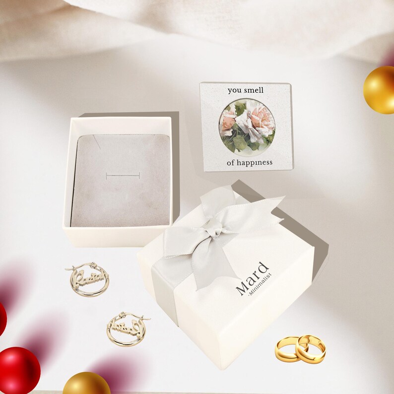Puede incluir: Vista a&eacute;rea de joyas y cajas de regalo. Dos pendientes de aro dorados con la palabra "Laila" est&aacute;n junto a una caja de joyer&iacute;a abierta. Tambi&eacute;n hay una caja de regalo blanca con una cinta plateada y la palabra "Mard". Tambi&eacute;n son visibles dos anillos de oro y una peque&ntilde;a tarjeta con flores.