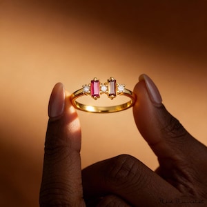 Op de afbeelding: Een gouden ring met twee rechthoekige edelstenen in roze en bruin, geflankeerd door kleine witte stenen. De ring wordt tussen twee vingers tegen een warme, neutrale achtergrond gehouden. Het ontwerp van de ring is elegant en modern.