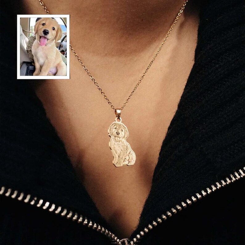 Puede incluir: Un collar dorado con un colgante con forma de perro. El colgante es una silueta detallada de un perro sentado. Una peque&ntilde;a foto enmarcada de un golden retriever est&aacute; en la esquina superior izquierda. El collar se lleva sobre un fondo negro.