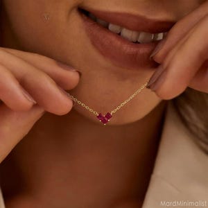 以下が含まれることがあります： 3つの正方形にカットされた深紅の宝石で構成された、ハート型のペンダントが付いた繊細なゴールドネックレス。 ネックレスは持ち上げられ、ペンダントのデザインとチェーンの細かいリンクが強調されています。 全体的な美しさはミニマリストでエレガントです。
