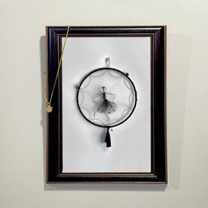 May include: A black wire sculpture of a ballerina is suspended inside a black wire dreamcatcher with a black tassel. The dreamcatcher is framed in a black wood frame with a gold chain hanging on the left side.