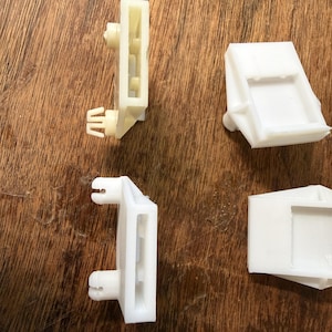 May include: Four white plastic automotive parts are displayed on a wooden surface. Two parts are rectangular with a slot and a protruding clip. The other two parts are square with a recessed area.