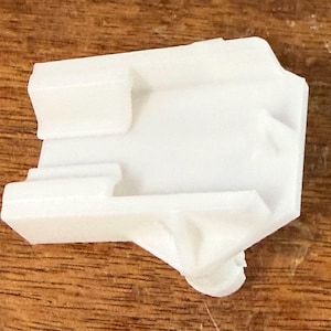May include: A white plastic component with a rectangular body and several protruding rectangular sections. The object has a rounded end and appears to be a part of a larger assembly. The component is set against a wooden surface.