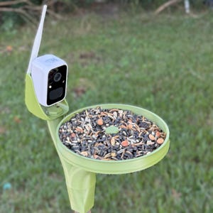 May include: A green bird feeder with a small camera attached to the side. The feeder is filled with birdseed and is mounted on a metal pole.