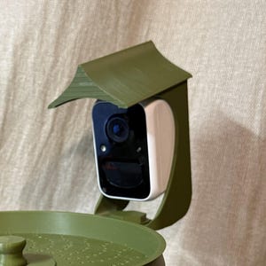 May include: A security camera with a white body and black face is mounted in a green, house-shaped enclosure. The camera has a lens and is designed to be mounted on a bird feeder. The enclosure has a small roof.