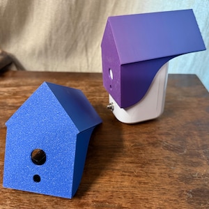 May include: Two decorative birdhouse-shaped objects. One is blue, the other is purple and attached to a white base. Both have circular openings. The objects are on a wooden surface.