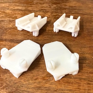 May include: Four white plastic components are arranged on a wooden surface. Two components have a rectangular shape with screw-like protrusions, and two have a more complex, multi-level design. The items appear to be 3D-printed.