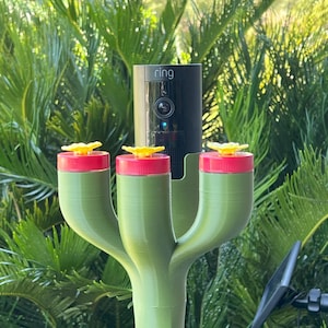 May include: A green, 3-pronged bird feeder with red and yellow accents. A black Ring doorbell camera is mounted on the top of the feeder.
