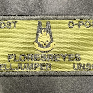May include: A rectangular patch with a black border and a light green background. The patch features the text "ODST", "O-POS", "FLORESREYES", "HELLJUMPER", and "UNSC". A black and yellow emblem is in the center.