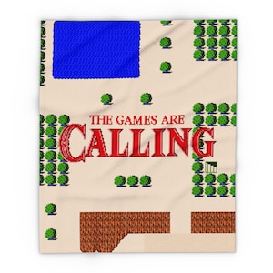 May include: A rectangular throw blanket featuring a pixelated 8-bit video game map design. The beige background displays a blue rectangular lake, green circular trees, and brown textured ground. The red text reads THE GAMES ARE CALLING.