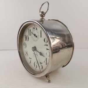 May include: A vintage silver-toned alarm clock with a round face and a small, circular clock face at the top. The clock has black numerals and hands on a white background. The clock is supported by three small feet and has a handle at the top.