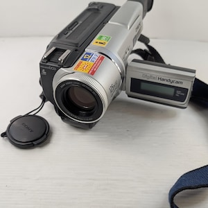 Peut inclure: Caméscope numérique Sony argenté et noir avec un objectif zoom optique 125x. L'appareil photo est équipé d'un bouchon d'objectif noir et d'une sangle bleue. L'écran affiche "Digital Handycam".