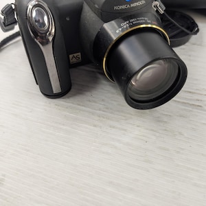 May include: Black Konica Minolta digital camera with a large lens and a silver accent. The camera has a strap and is next to a black carrying case. The camera has the text "KONICA MINOLTA" on the top.