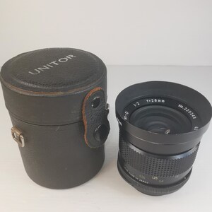 Peut inclure: Un objectif d'appareil photo noir avec une bague argentée et le texte "HI-D 1:2 f=28mm 030 MADE IN JAPAN 125 04 No.220248" imprimé dessus. L'objectif est dans un étui en cuir noir avec le mot "UNITOR" imprimé dessus.