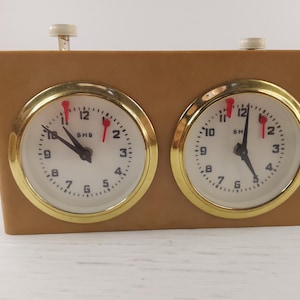 Può includere: Un orologio da scacchi vintage con cassa marrone e due quadranti rotondi. Ogni quadrante ha uno sfondo bianco, numeri neri e finiture dorate. Le lancette sono nere e la lancetta dei secondi è rossa.