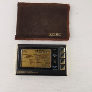 Può includere: Una sveglia Seiko al quarzo con orario mondiale vintage con cornice color oro e quadrante nero. Il display mostra l'ora, la data e i fusi orari mondiali. È inclusa anche una custodia protettiva marrone con il logo Seiko.