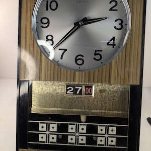 May include: A vintage Seiko wall clock with a silver face, black hands, and a wood-effect frame. The clock displays the day and date, with buttons below the clock face. The rectangular clock has a retro design, reminiscent of mid-century decor.
