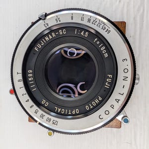 Peut inclure: Un objectif de caméra Fujinar-SC vintage avec un obturateur Copal No.3. L'objectif a un boîtier en métal argenté et noir avec des marquages dont "1:4.5 f=18cm" et "FUJI PHOTO OPTICAL CO." L'objectif est monté sur une base en bois.