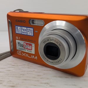 May include: Orange Casio Exilim digital camera with a silver lens and a grey strap. The camera has a YouTube capture mode and 8.1 megapixels. The lens is 7.4-18.6mm.