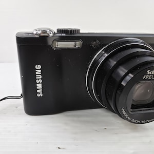 May include: A black Samsung digital camera featuring a Schneider Kreuznach Varioplan Zoom 4.0-72mm 1:3.2-5.8 24mm lens. The camera body includes a built-in flash, a silver shutter button, a black wrist strap, and the Samsung logo printed on the front.