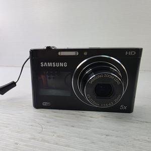 Könnte beinhalten: Eine schwarze Samsung Digitalkamera mit einem 5-fach Zoomobjektiv und den Spezifikationen 4.5-22.5mm 1:2.5-6.3 25mm. Die Vorderseite zeigt das Samsung-Logo, ein WLAN-Symbol, eine HD-Kennzeichnung und eine schwarze Handschlaufe am Kameragehäuse.