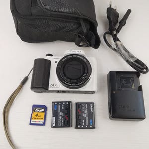 Pode incluir: Uma câmera digital Casio Exilim branca com punho e lente pretos, zoom 24x. Inclui uma capa preta, carregador, duas baterias, um cartão SD e um cabo de alimentação. A câmera tem uma alça de pulso.