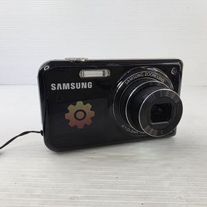 Puede incluir: Una cámara digital Samsung negra con un objetivo Samsung Zoom Lens con las especificaciones de 4,7-23,5 mm impresas en la parte frontal. El cuerpo de la cámara muestra el logotipo de Samsung, un icono de engranaje y una correa negra.