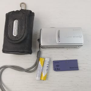 May include: A silver Sony Cyber-shot U digital camera with a black case, two AA batteries, and a Memory Stick. The camera has a wrist strap and the case has a metal clasp.