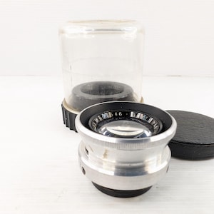 Peut inclure: Un objectif d'appareil photo vintage avec un corps argenté et des détails noirs, accompagné d'un étui en plastique transparent et d'un bouchon d'objectif noir. L'objectif comporte des marquages visibles.