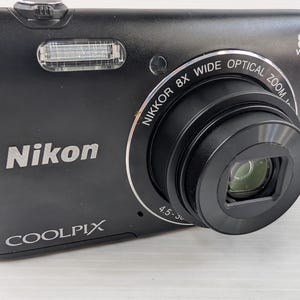 May include: A black Nikon Coolpix digital camera featuring an 8x wide optical zoom lens. The front of the camera displays the Nikon logo, the Coolpix model name, and text around the lens barrel reading Nikkor 8x wide optical zoom 4.5-36mm.