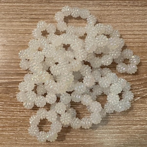 Peut inclure: Plusieurs élastiques à cheveux blancs et irisés en perles, disposés sur une surface en bois. Les élastiques sont circulaires avec un motif texturé en forme d'étoile. Les perles scintillent et captent la lumière.