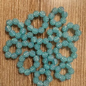 Peut inclure: Une collection d'ornements circulaires bleus clair, perlés. Chaque ornement est composé de petites perles texturées disposées en forme d'anneau. Les ornements sont dispersés sur une surface en bois.