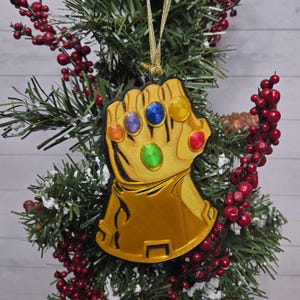 May include: A gold-colored Thanos Infinity Gauntlet Christmas ornament with six colorful gems. The ornament is hanging on a Christmas tree with red berries and green needles. The gauntlet is detailed with black outlines and a gold string for hanging.