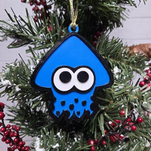 May include: A blue and black squid-shaped Christmas ornament with white eyes and a gold string for hanging. The ornament is hanging on a Christmas tree with red berries and green needles.
