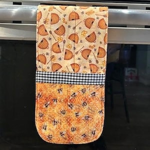 May include: A quilted oven mitt with a bee and honeycomb design. The top section features a pattern of honeycombs and spoons on a beige background. The bottom section has a pattern of bees on an orange background, separated by a black and white checkered band.