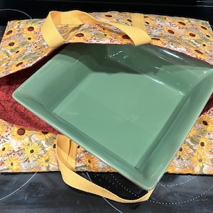 May include: A green square baking dish rests inside a fabric carrier. The carrier has a yellow sunflower pattern on a cream background, with a burgundy lining. Yellow straps are attached to the carrier.