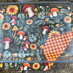 May include: A clear glass baking dish with a colorful, patterned insert featuring mushrooms, sunflowers, and foliage. A folded orange and red plaid fabric is visible. The dish is on a wooden surface.