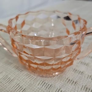 May include: A vintage pink glass sugar bowl with two handles. The bowl has a diamond-cut pattern and a scalloped rim. The sugar bowl is made of clear glass and has a light pink hue. It is a decorative piece for serving sugar or other small items.